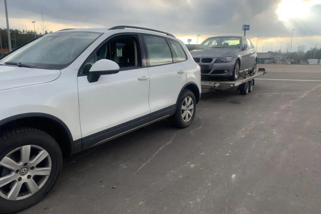 Weißer Geländewagen mit Autotransport-Anhänger, darauf ein grauer BMW Kombi, auf einem großen Parkplatz im Abendlicht