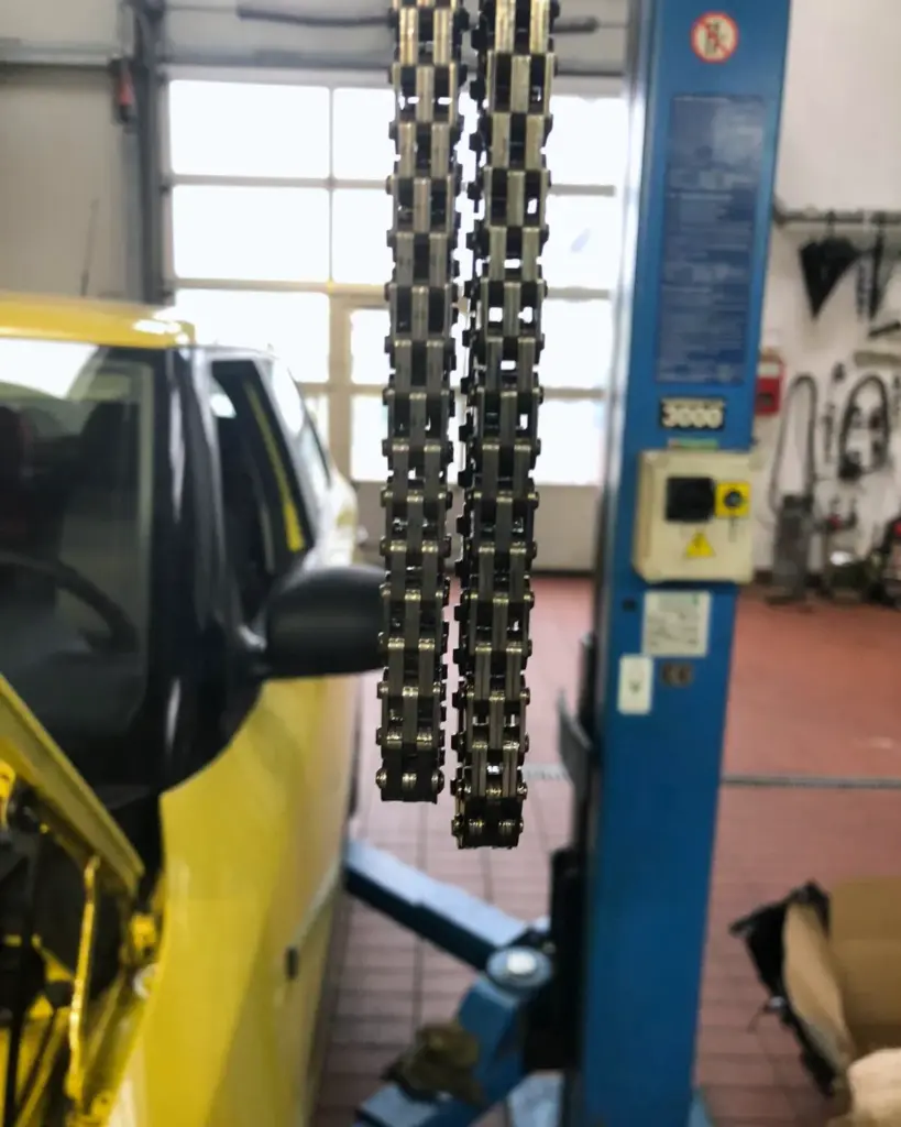 Zwei ausgebaute Steuerketten hängen nebeneinander vor der Hebebühne, links ein gelbes Auto